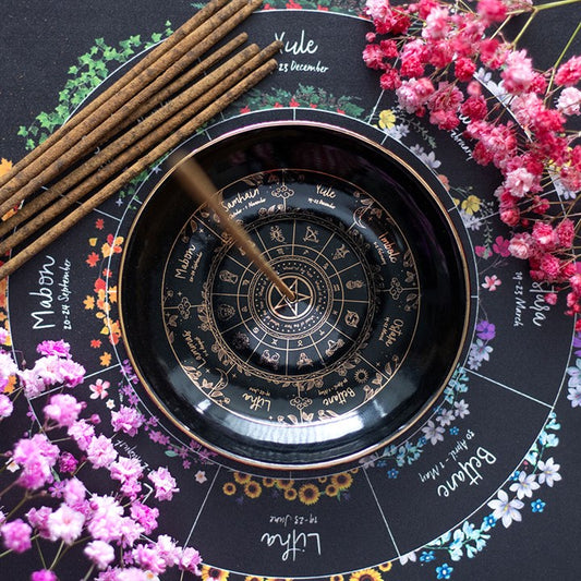 A black Wheel of the Year divination wheel featuring zodiac signs and pagan holidays, surrounded by pink and purple dried flowers, incense sticks, and a spinning pendulum—perfect for meditation spaces.