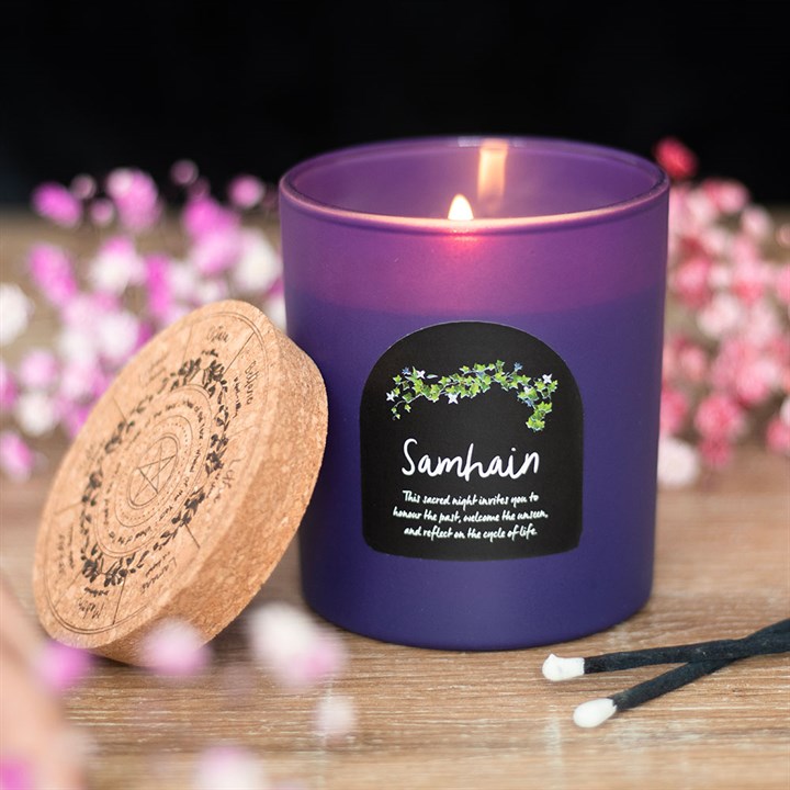 A lit purple candle labelled “Samhain” sits beside a cork lid adorned with a pentagram and botanical designs. Pink and white flowers, along with two black matches, surround this perfect addition to autumn altars on a wooden surface.