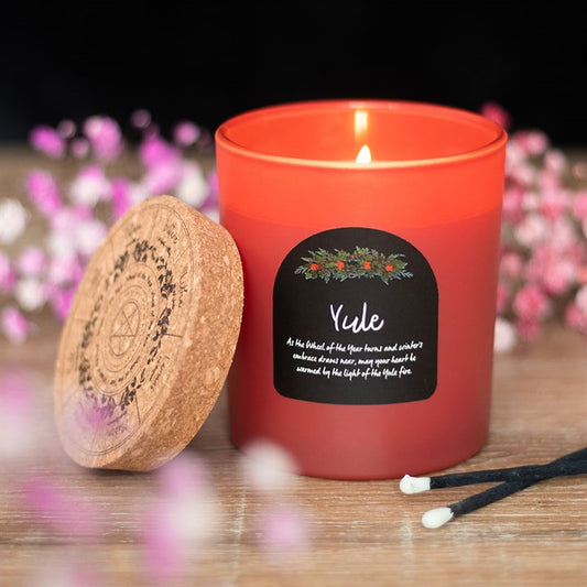 A red Yule candle labelled “Yule” with a floral design burns beside a cork lid engraved with a pentacle, crafted from vegan paraffin wax and surrounded by black-tipped matches and blurred pink and white flowers.