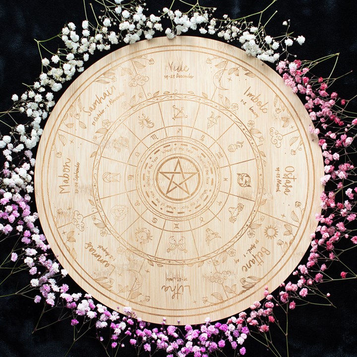 A circular wooden Wheel of the Year, engraved with zodiac signs, seasonal symbols, and a pentacle at its centre, is surrounded by a gradient ring of baby’s breath flowers that fade from white to pink against a black background.