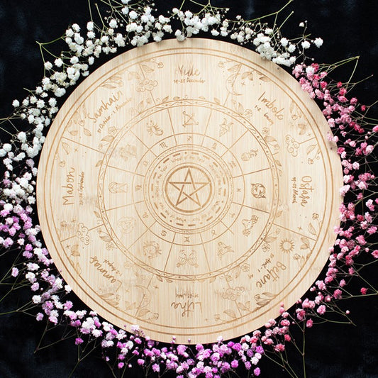 A circular wooden Wheel of the Year, engraved with zodiac signs, seasonal symbols, and a pentacle at its centre, is surrounded by a gradient ring of baby’s breath flowers that fade from white to pink against a black background.