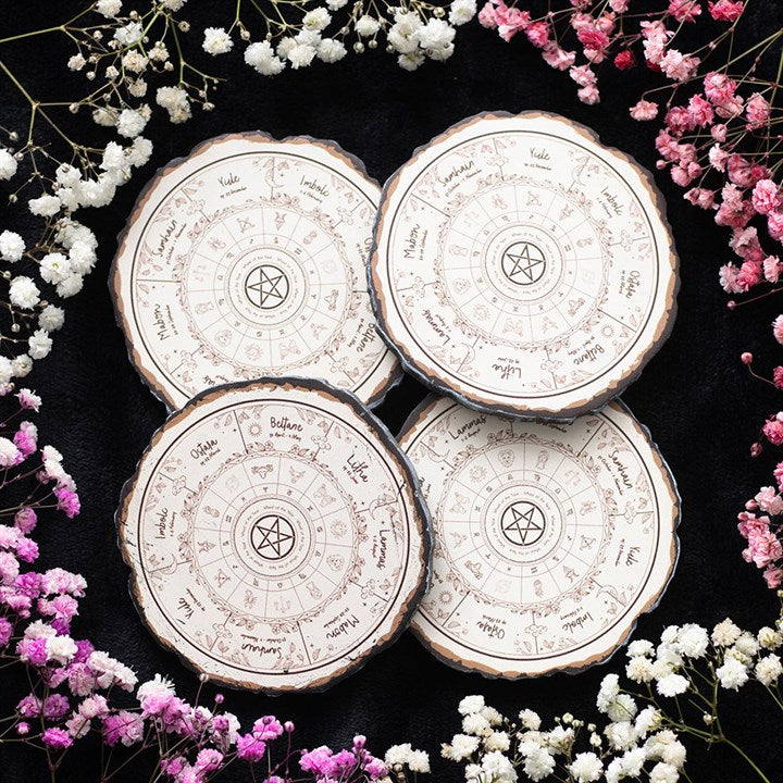 Four round wooden slice coasters engraved with zodiac charts and pentagram symbols are arranged in a circle on a black background—perfect for spiritual homes—and surrounded by clusters of white and pink babys-breath flowers.
