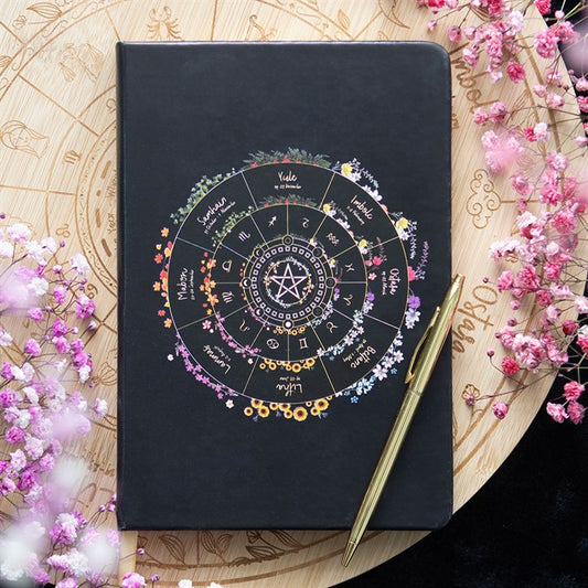 A black notebook with a colourful astrological wheel design, perfect for journalling or seasonal reflections, rests on a wooden zodiac chart engraved with astrology symbols. Pink and white flowers are scattered around.