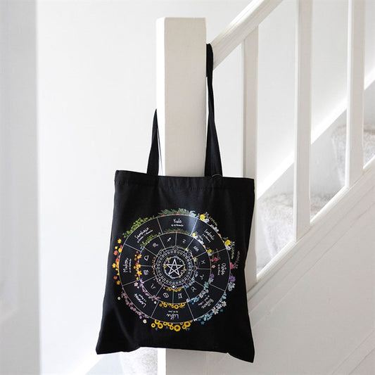 A black polycotton tote bag with a colourful Wheel of the Year design hangs on a white stair bannister, featuring astrological symbols, names, and a pentagram at the centre, surrounded by floral accents.