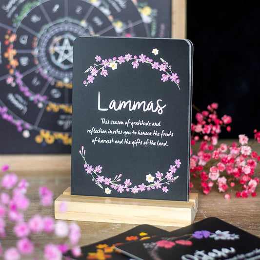 A card on a wooden stand displays “Lammas” in white script, surrounded by pink and purple flowers. Perfect for seasonal altars, it honours the fruits of the harvest as part of the Wheel of the Year. Pink flowers and a zodiac chart are visible in the background.