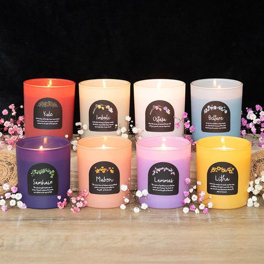 Eight colourful scented candles in glass jars, each labelled with a Wheel of the Year celebration, are lit and arranged on wood. Pink and white flowers decorate the ritual gift set against a black background.