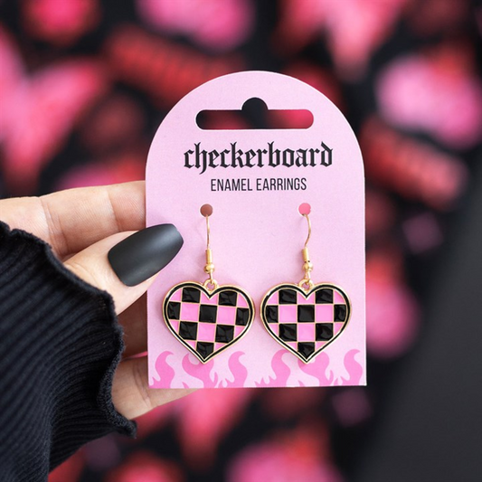 A hand with black matte nails holds a pink card labelled checkerboard ENAMEL EARRINGS, displaying heart-shaped earrings in an alternative style with a pink and black chequerboard pattern and gold accents. Blurred pink shapes appear behind.