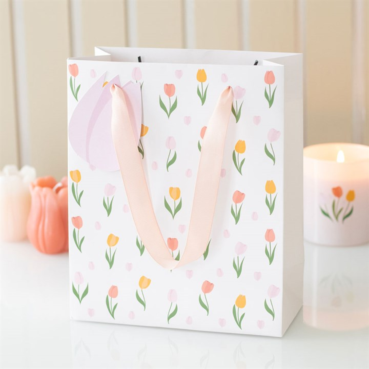 A white floral gift bag with pink handles displays pastel tulips in pink, yellow, and orange. It rests on a white surface beside a lit candle and a tulip-shaped candle, with a beige striped wall behind.