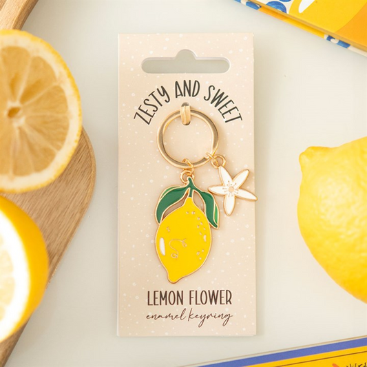 A citrus charm with a bright yellow enamel lemon and white flower dangles from this lemon flower keyring, fastened to a card that says "Zesty and Sweet," surrounded by fresh lemons and slices.