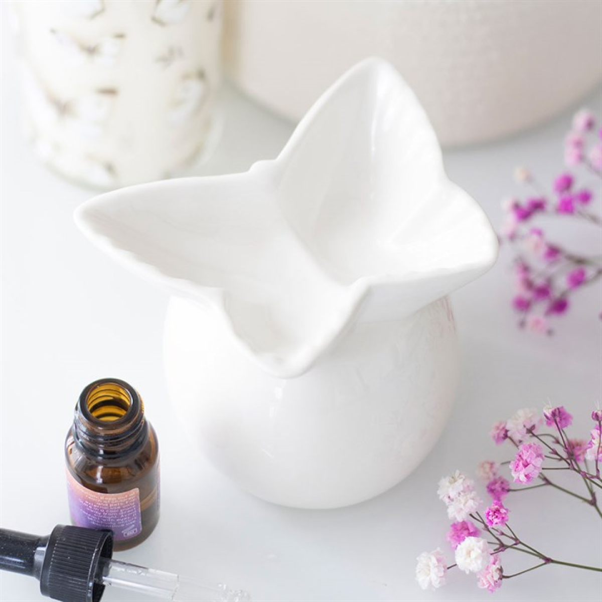 A white ceramic oil burner in the shape of a butterfly rests on a table, accompanied by a brown fragrance oil bottle, a dropper, delicate pink flowers, and a white container in the background.