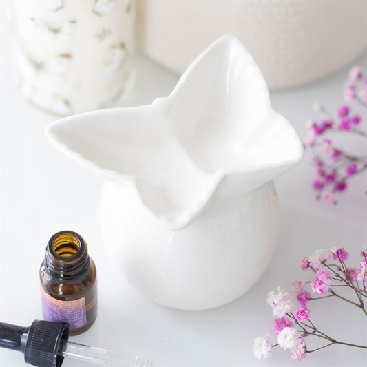 A white ceramic oil burner in the shape of a butterfly rests on a table, accompanied by a brown fragrance oil bottle, a dropper, delicate pink flowers, and a white container in the background.