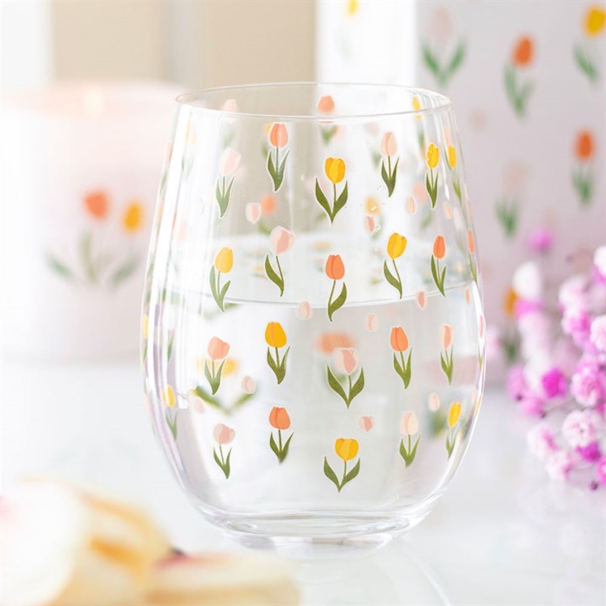 A clear, stemless glass adorned with small tulip patterns in pink, yellow and peach rests on a white surface. Blurred pink flowers and decorative items appear in the background—ideal for Mother’s Day.