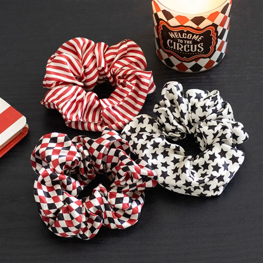 Three circus scrunchies with vintage star prints—red and white stripes, red and white diamonds, and black stars on white—rest on a black surface beside a candle labelled “Welcome to the Circus.”.