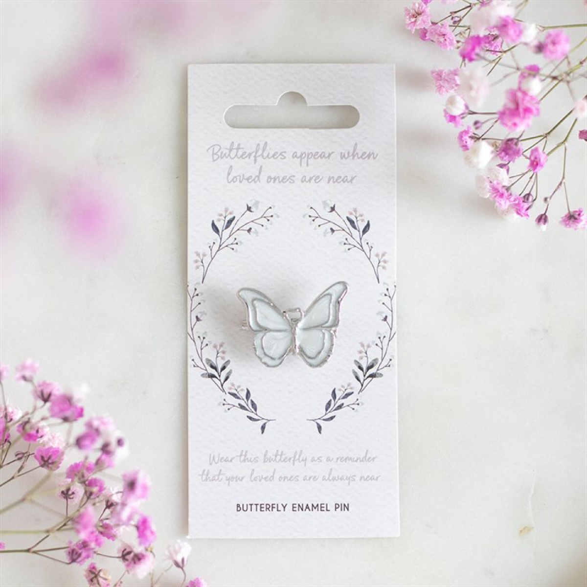 A white enamel butterfly pin is fastened to a card with a delicate leaf wreath design. The card reads: “Butterflies appear when loved ones are near.” Pink baby's breath flowers frame this remembrance gift.