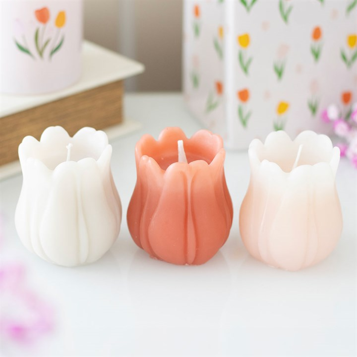 Three tulip-shaped candles—white, coral pink, and pale pink—are placed side by side on a white surface, ideal for spring table settings. In the background, floral-patterned containers and blurred pink flowers appear.