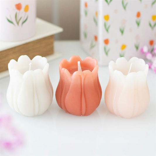 Three tulip-shaped candles—white, coral pink, and pale pink—are placed side by side on a white surface, ideal for spring table settings. In the background, floral-patterned containers and blurred pink flowers appear.