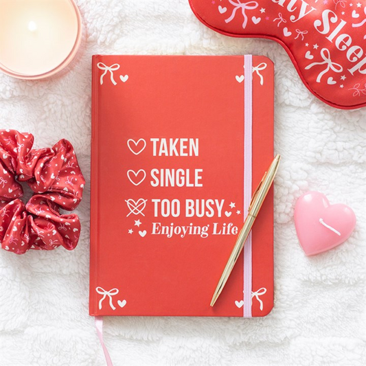 A red notebook with lined pages and a pink elastic band lies on a white textured blanket, displaying “TAKEN, SINGLE, TOO BUSY ENJOYING LIFE,” surrounded by candles, a scrunchie, and a red sleep mask.