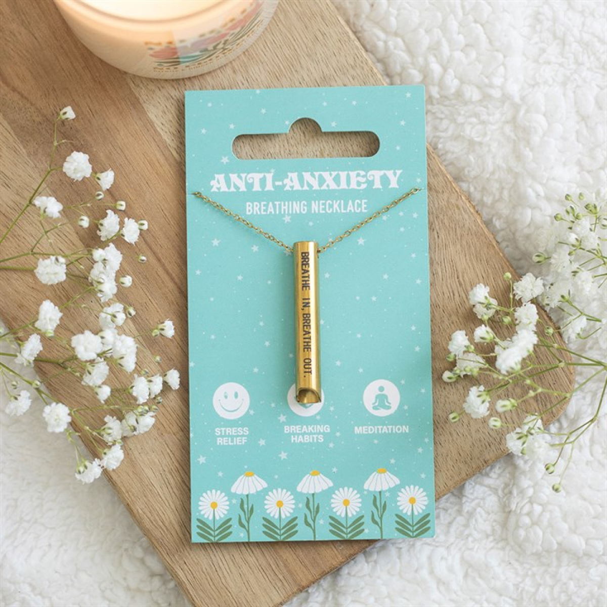 A gold anti-anxiety breathing necklace inscribed “Breathe In. Breathe Out.” sits on teal packaging, encircled by white flowers and a candle—elegant stress-relief jewellery for mindful moments.