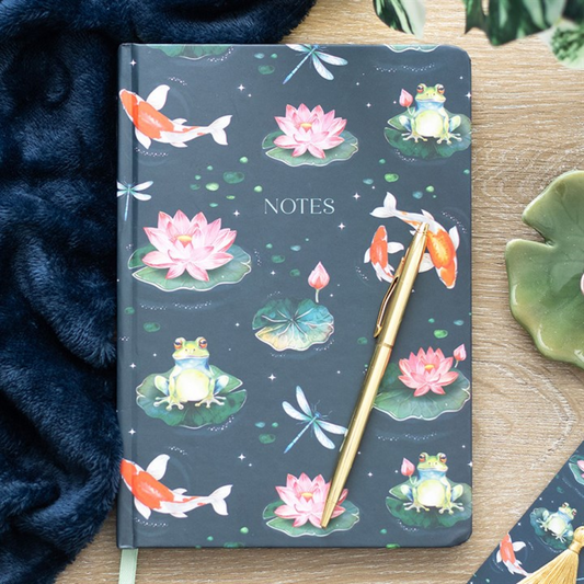 A pond-themed A5 notebook with a dark cover features koi carp, pink water lilies, lily pads, a frog and dragonflies. A gold pen lies diagonally on top. The lined notebook is on a wooden surface near dark blue fabric.