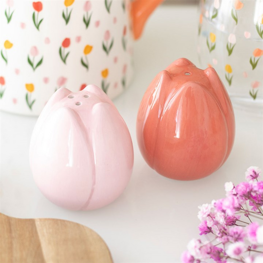 Two ceramic salt and pepper shakers shaped like tulip buds—one light pink, one coral—rest on a white surface, surrounded by floral kitchen décor including pink flowers and a white jug with a tulip design.