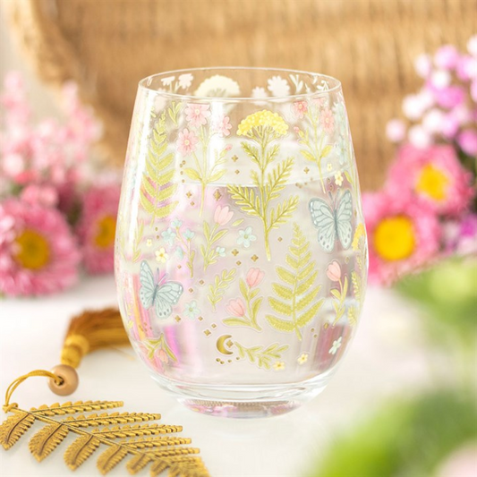 A clear, stemless glass adorned with a pastel floral design, green leaves, and blue butterflies. Gold accents enhance the butterfly motifs. Blurred pink flowers and a gold fern tassel create a soft, floral setting.