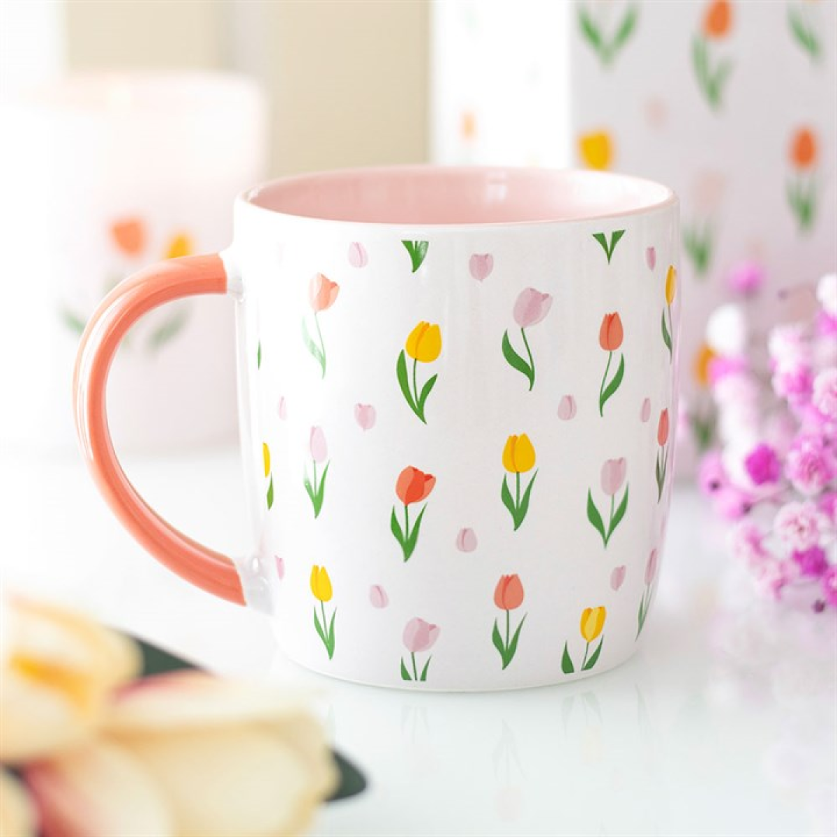 A white ceramic mug with a pink handle and interior displays a delightful tulip print. The mug sits on a white surface surrounded by blurred pink flowers and soft pastel items in the background.
