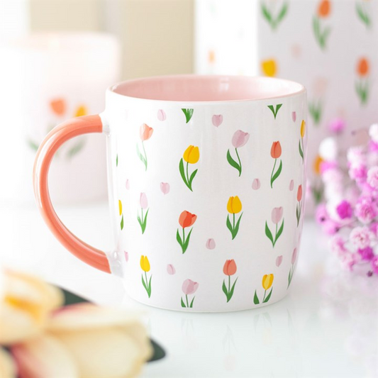 A white ceramic mug with a pink handle and interior displays a delightful tulip print. The mug sits on a white surface surrounded by blurred pink flowers and soft pastel items in the background.