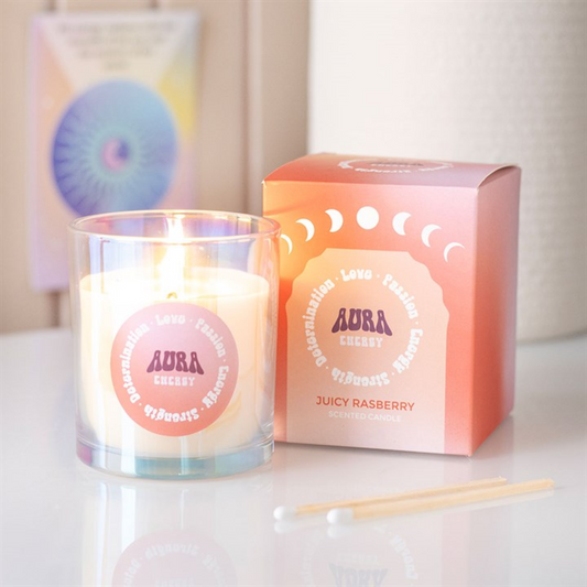 A lit iridescent glass candle labelled “AURA ENERGY” rests beside its pink box marked “Juicy Raspberry Scented Candle.” Two wooden matches and a cotton bud are nearby on a white surface. A colourful card is blurred in the background.