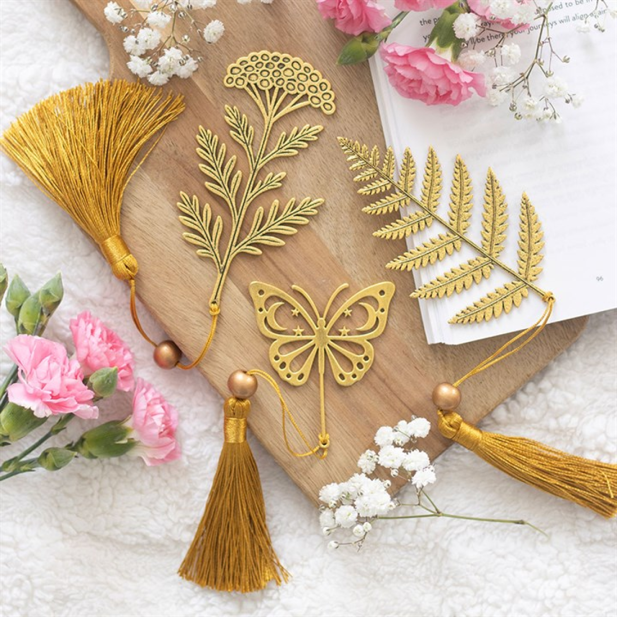 Four golden metal bookmarks shaped like leaves, a flower, and a butterfly with yellow tassels rest on a wooden board, ideal as nature-themed bookmarks or thoughtful bookshop gifts for any reader.