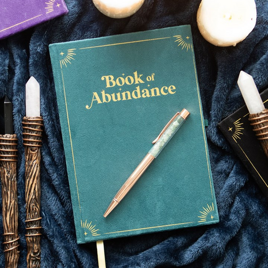 A green Book of Abundance journal with gold accents sits on dark blue fabric, surrounded by a green aventurine crystal pen, candles, and decorative wands embellished with white and black crystals.