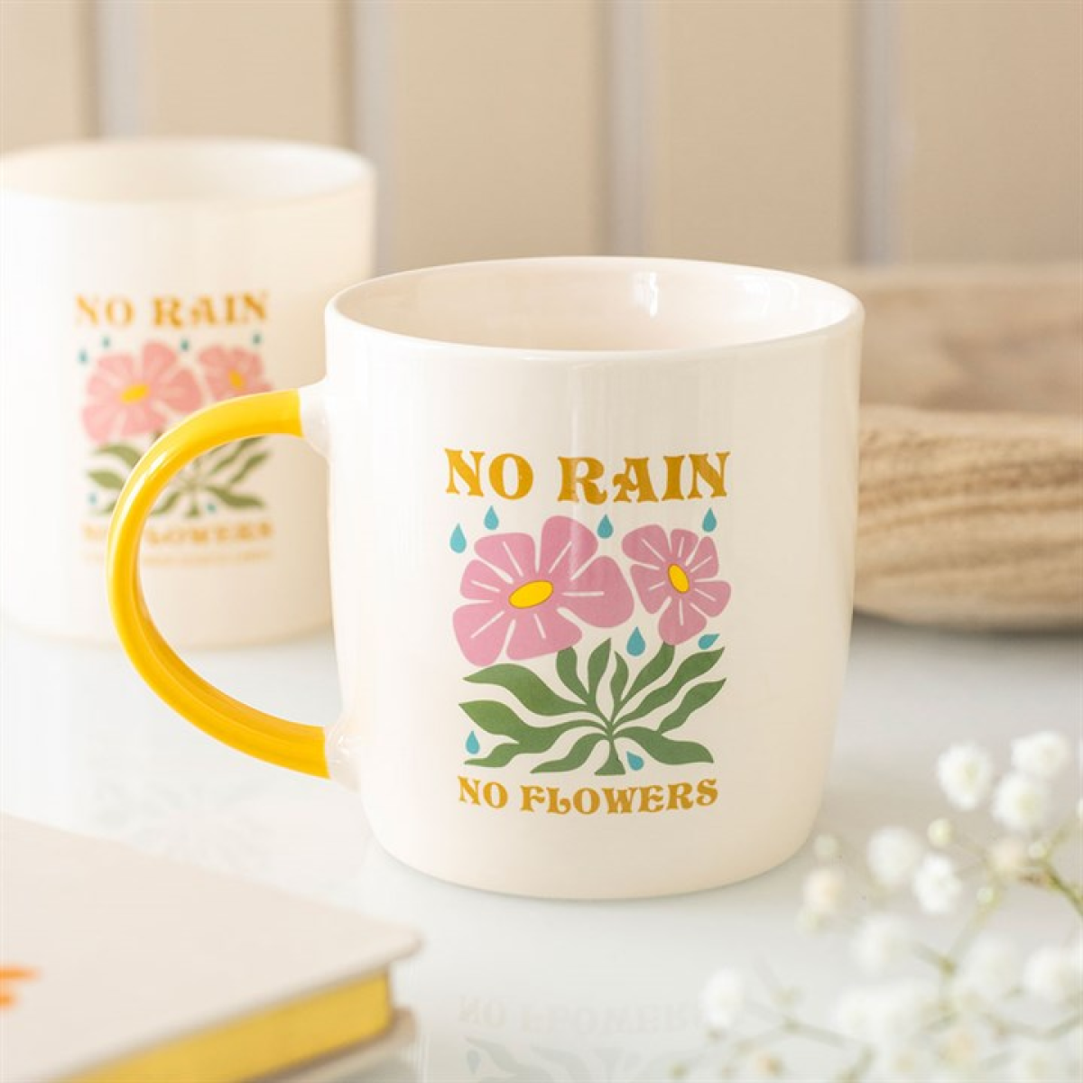 A cheerful mug with a yellow handle displays pink flowers, green leaves, and the words “NO RAIN NO FLOWERS” in yellow—an uplifting gift, with another similar No Rain No Flowers mug blurred in the background.