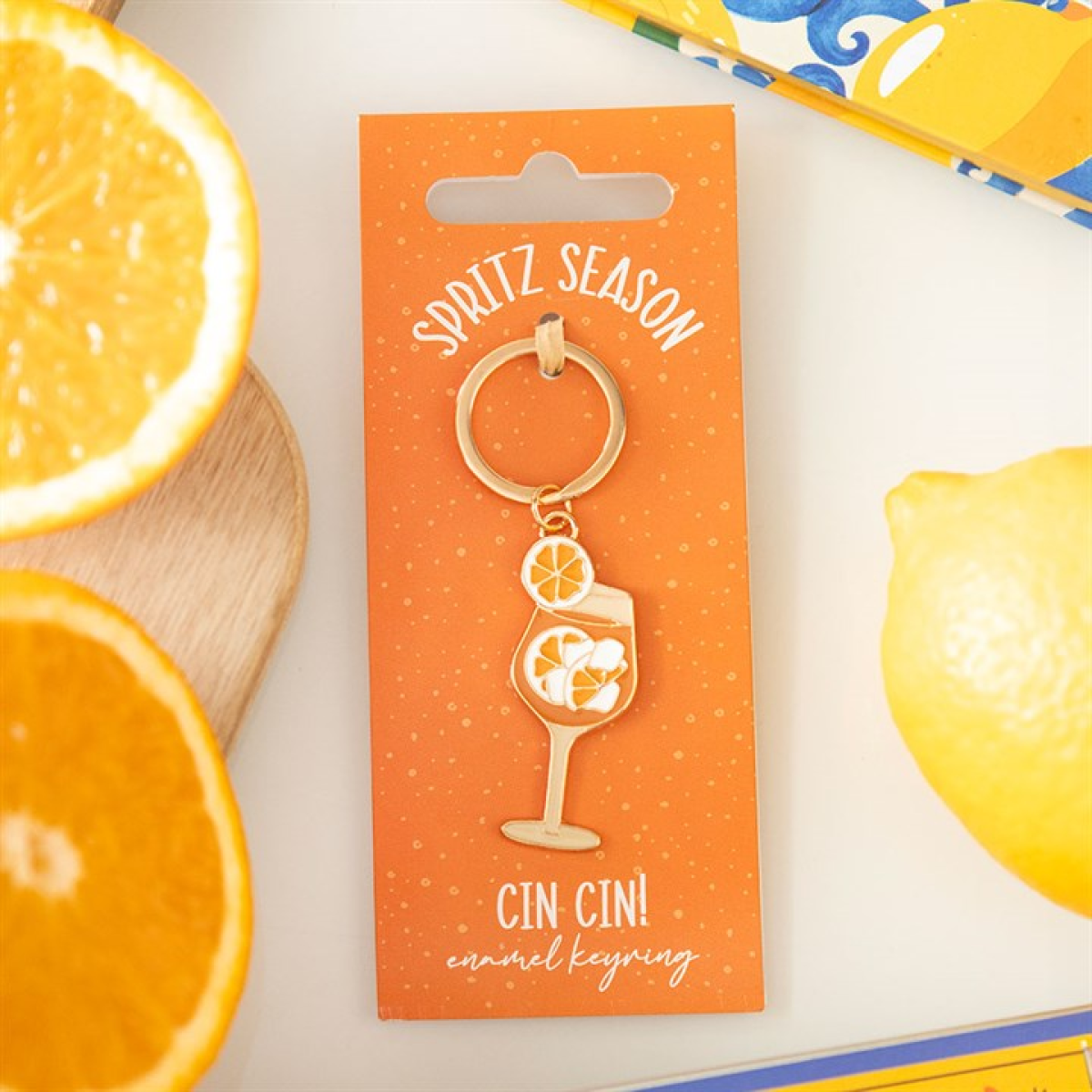A gold enamel keyring in the shape of a spritz cocktail with orange slices, attached to an orange card reading 'Spritz Season'—ideal as a summer accessory—surrounded by orange and lemon halves.