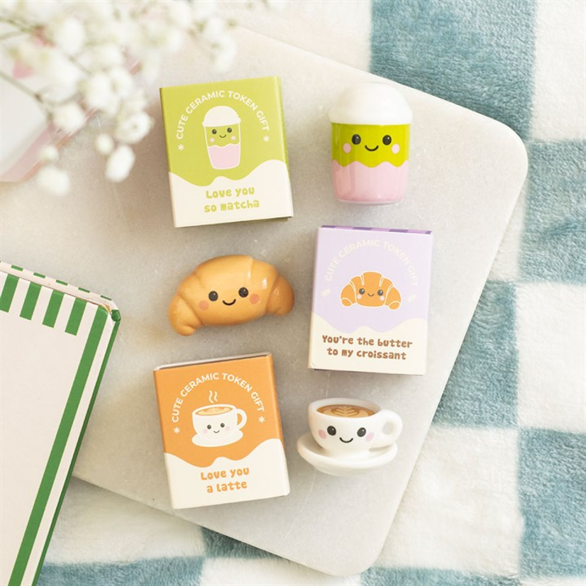 Four small boxes feature puns and kawaii ceramic tokens—charming café mascot gifts such as a matcha drink, croissant, and latte with smiling faces—arranged on a chequered blanket beside a green-striped notebook and white flowers.
