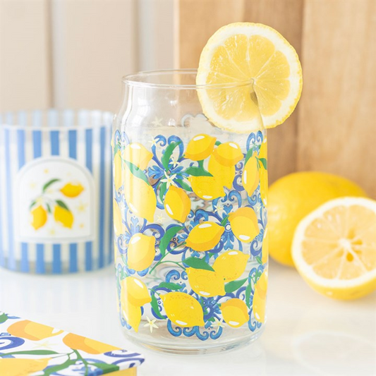 A can-style glass adorned with vivid yellow lemons and blue swirls, filled with a clear liquid and topped with a lemon slice. Ideal summer tableware, encircled by whole lemons and a blue-striped candle.