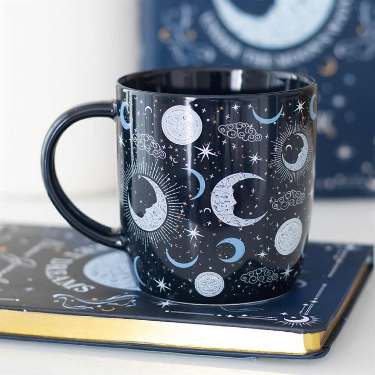 A black ceramic blue moon mug with silver and blue celestial designs rests atop a closed journal with gold-edged pages. This 500ml microwave-safe mug suits stargazers, with a matching celestial-themed book in the background.