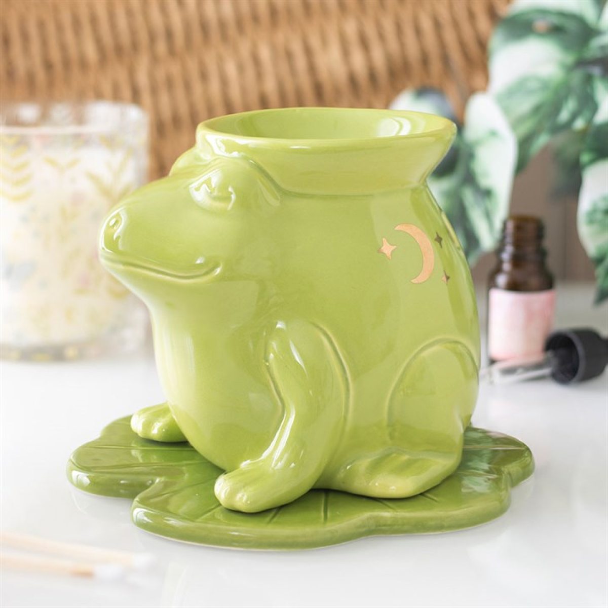 A glossy green frog oil burner, adorned with crescent moon and star shapes, rests on a matching lily pad. In the background are plants, a dropper bottle, and a glass candle holder for home fragrance.