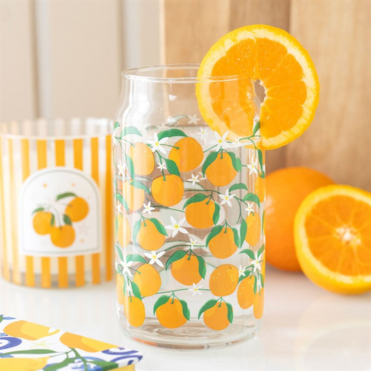 A clear can-style glass with orange print and white flowers, filled with water and garnished with a fresh orange slice. In the background are an orange candle in a striped holder and two halved oranges on a white surface.