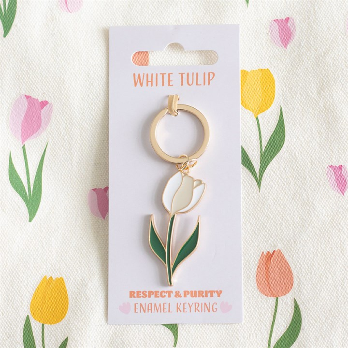 A charming keyring with a gold and white tulip design hangs from a card marked WHITE TULIP, set against pastel floral artwork. Beneath, the words RESPECT & PURITY ENAMEL KEYRING are displayed.