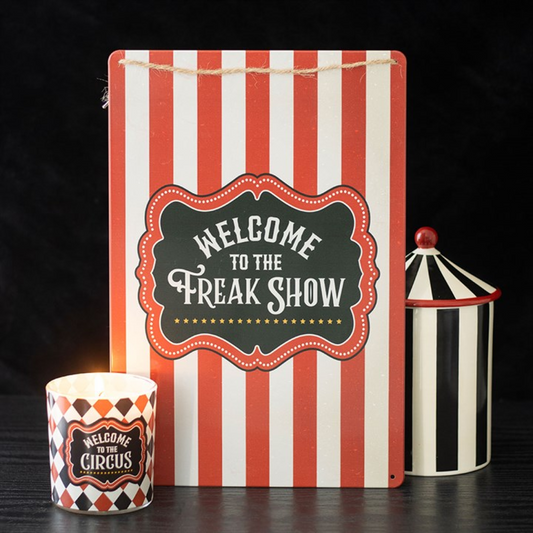 A metal hanging sign with red and white vertical stripes reads 'Welcome to the Freak Show' in vintage-style lettering. In front is a lit candle marked 'Welcome to the Circus', with a striped container—ideal freak show décor.