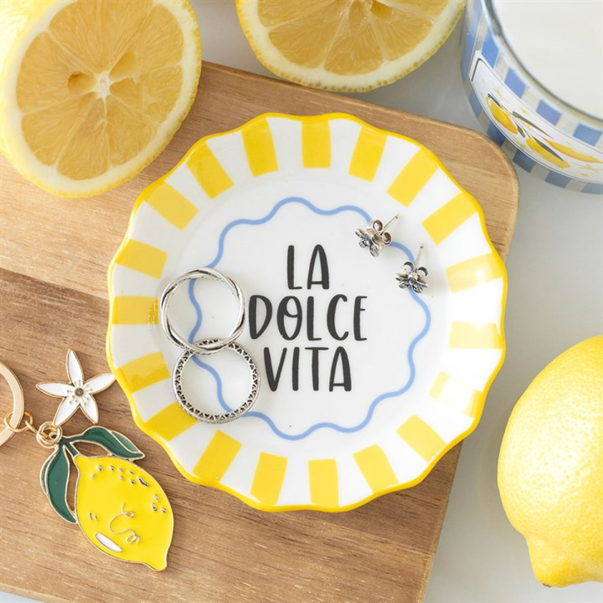 A ceramic trinket dish with yellow stripes and “LA DOLCE VITA” in the centre holds two rings and bee earrings, surrounded by sliced lemons, a keyring, and a lemon-themed mug on a bedside table.