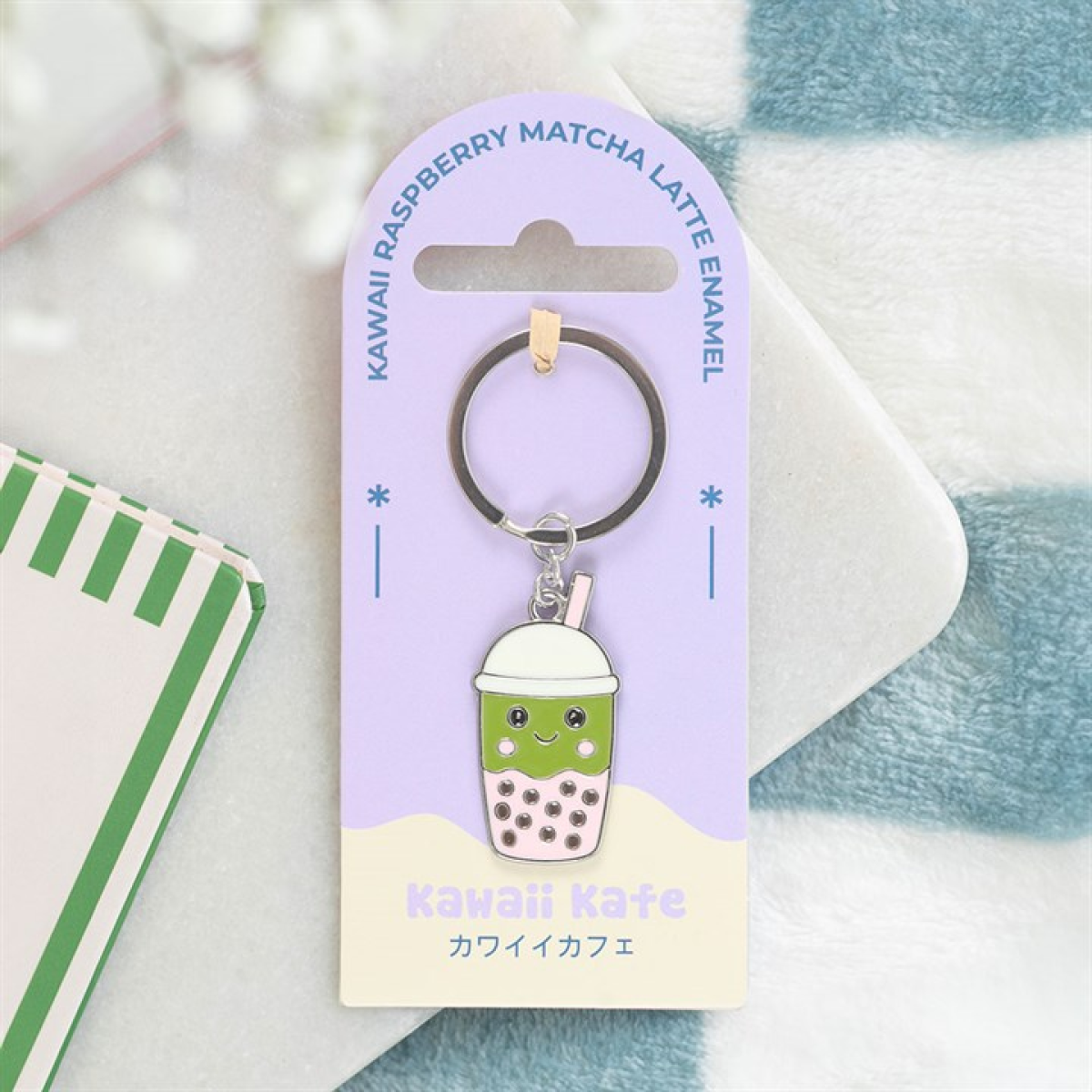 A charming green bubble tea accessory keyring with a smiling face is attached to a lavender card marked “Kawaii Raspberry Matcha Latte Enamel.” The card also says “Kawaii Kafe” in both English and Japanese.