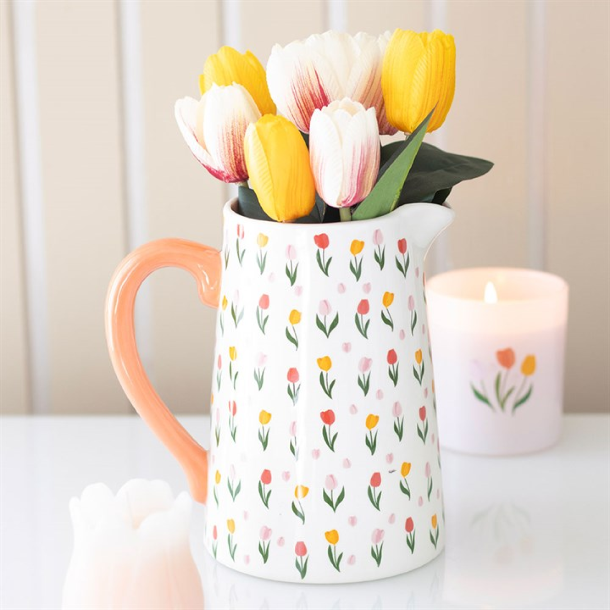 A white ceramic flower jug adorned with small tulip designs contains yellow and white tulips. Nearby, a lit candle with a tulip motif rests on a white surface against a beige panelled background. Ideal Mother’s Day gift.