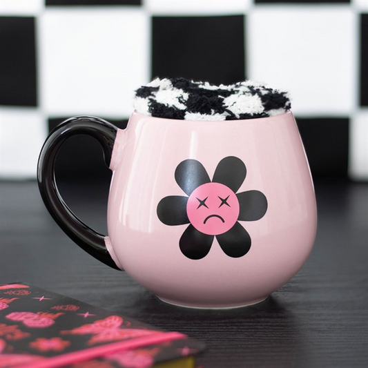 A pink mug with a sad flower and black handle holds fluffy chequerboard socks. A pink notebook sits nearby against a black-and-white chequerboard background—an ideal emo present.