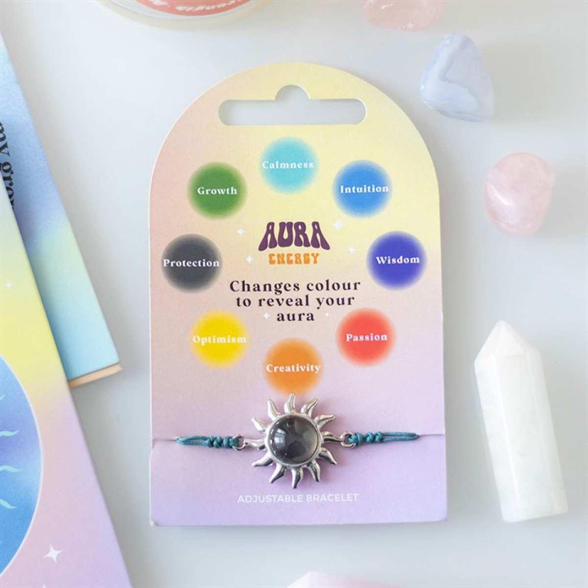 A sun charm colour-changing bracelet with a dark centre rests on a pastel “Aura Energy” card, displaying mood circles such as Growth and Passion, and promises to reveal your aura as the bracelet’s colour changes.
