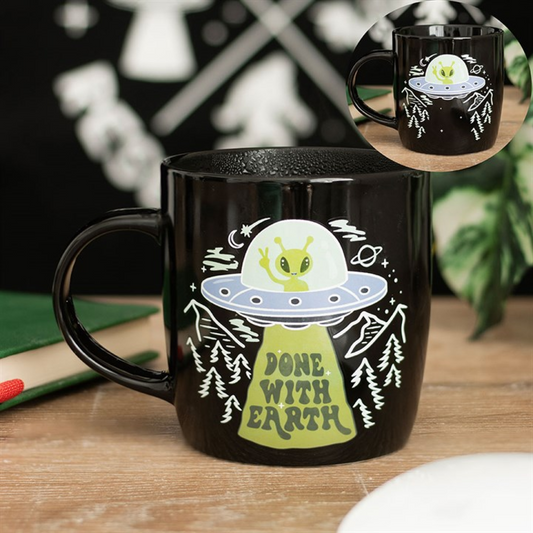 A novelty coffee mug featuring a cartoon green alien in a UFO, beaming up trees. The beam reads DONE WITH EARTH in green lettering. The alien mug rests on a wooden table next to a green notebook and white plate.