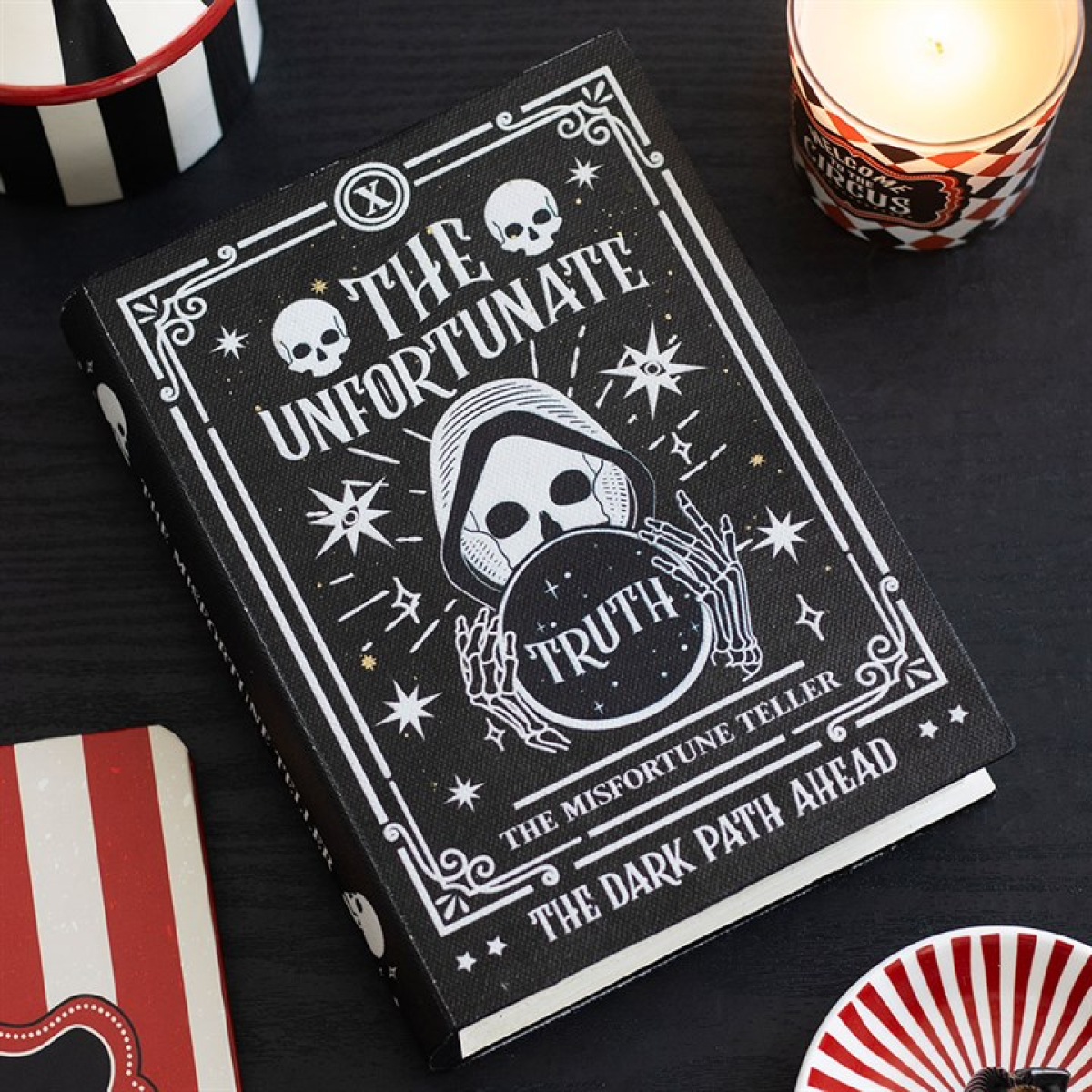 A black book titled The Unfortunate Truth shows a hooded, skeletal fortune teller holding a crystal ball. The cover displays skulls, stars, and ornate borders with tarot cards and circus-themed items nearby on a dark surface.