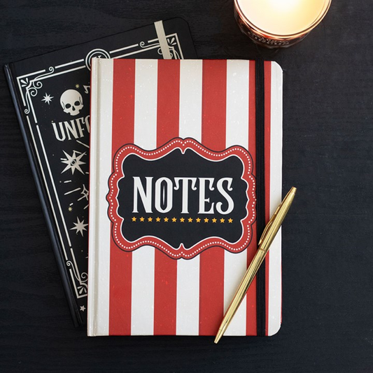 A vintage circus notebook with red and white stripes marked “NOTES”, along with a black and gold pen, rests atop a black skull-and-stars lined journal beside a lit candle on a dark surface.