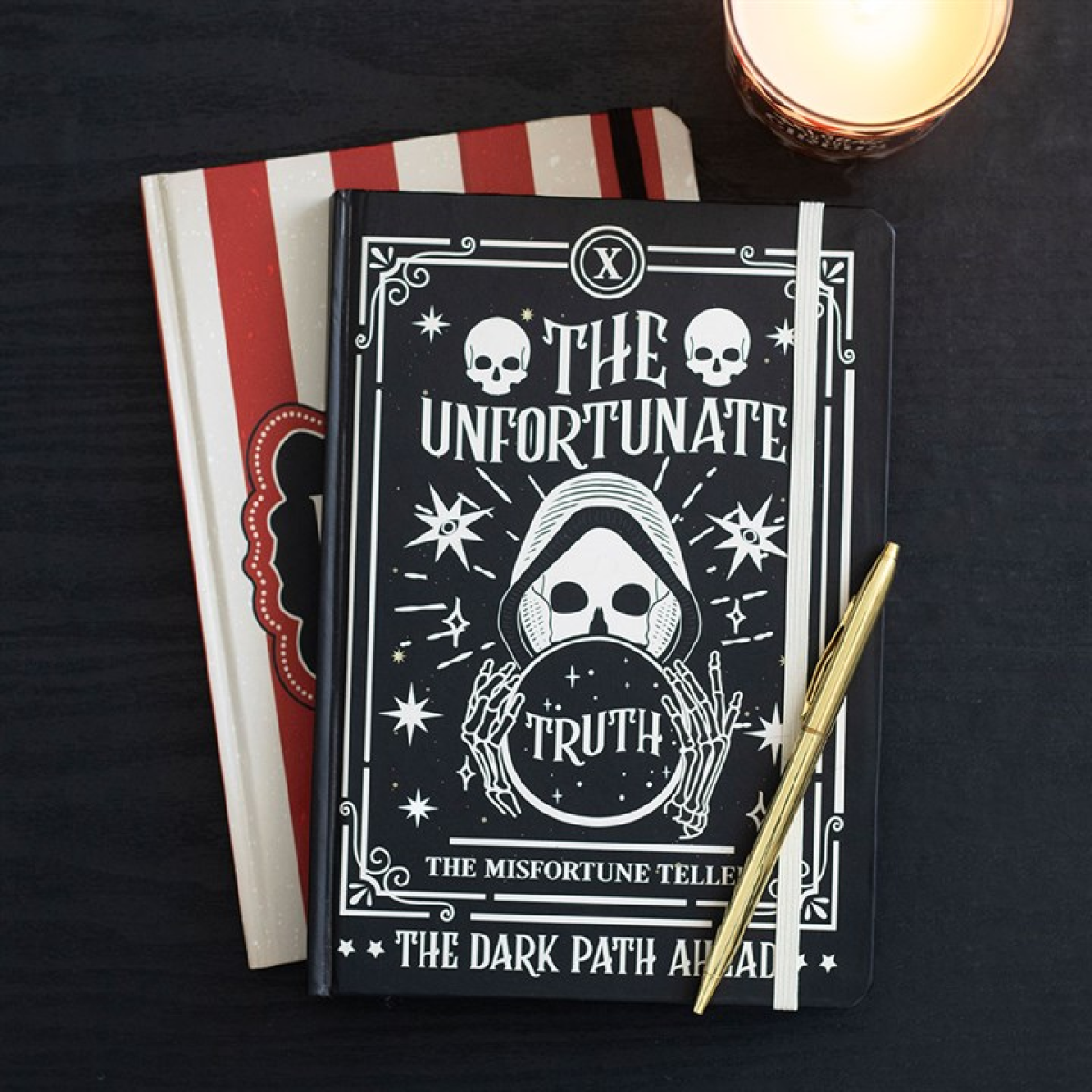 A black fortune teller’s notebook featuring a mystical skull and hood design bears the words “The Unfortunate Truth” and “The Dark Path Ahead.” It sits on a red and white striped book near a lit candle and gold pen on a dark surface.