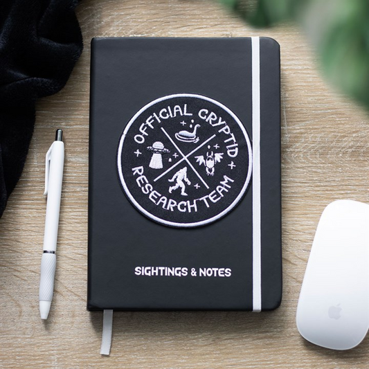 A black cryptid notebook labelled “SIGHTINGS & NOTES,” featuring an “Official Cryptid Research Team” patch, rests on a wooden desk beside a white pen and an Apple Magic Mouse.
