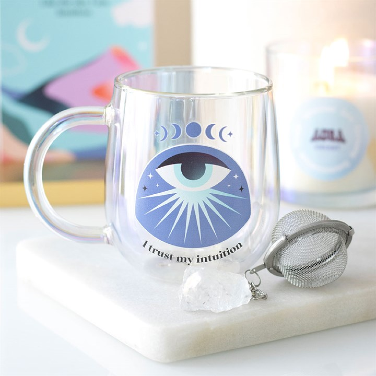 A clear mug with an all-seeing eye design features crescent moons, stars, and the phrase “I trust my intuition.” Nearby are a crystal tea infuser, a white crystal, and a lit candle on a white surface.
